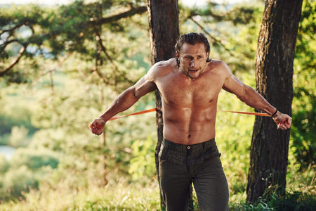 Doing workout. Handsome shirtless man with muscular body type is in the forest at daytime.