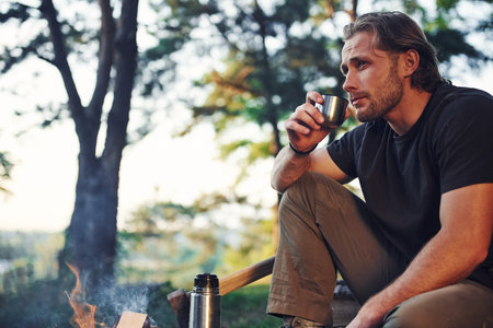Making drink by using thermos. Man in black shirt near the campfire in the forest at his weekend...