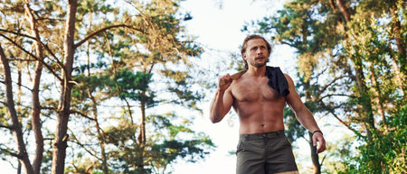 With axe. Handsome shirtless man with muscular body type is in the forest at daytime.