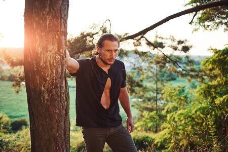 Leaning on the tree. Amazing sunlight. Handsome man with muscular body type is in the forest at daytime.の写真素材