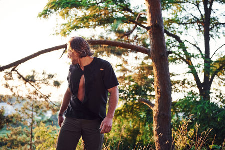 Looks behind. Handsome man with muscular body type is in the forest at daytime.