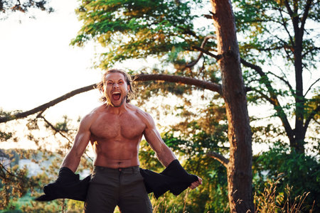 Looks behind. Handsome shirtless man with muscular body type is in the forest at daytime.