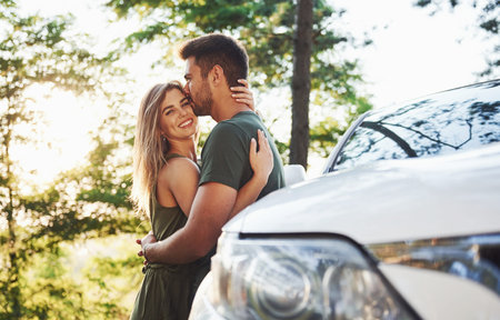 Front part of white car. Beautiful young couple have a good time in the forest at daytime.