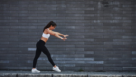 Young sportive brunette with slim body shape against brick wall in the city at daytime.