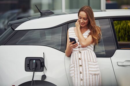 Using smartphone while waiting. Woman on the electric cars charge station at daytime. Brand new vehicle.の写真素材