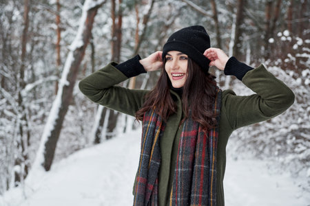 Touches the black hat. Cheerful young girl in warm clothes have a walk in the winter forest at...