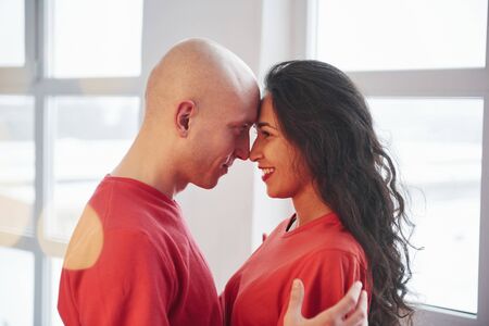 Near the windows. Lovely couple in red clothes standing in the white room and looking at each other.の写真素材