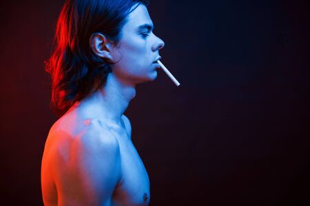 Guy smokes a cigarette. Studio shot in dark studio with neon light. Portrait of serious man.の写真素材