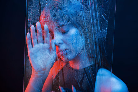 Feel tired. Studio shot in dark studio with neon light. Portrait of beautiful girl behind wet glass.の写真素材