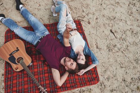 Acoustic guitar near them. Young couple have picnic on the beach. Lying on the red colored blanket.の写真素材