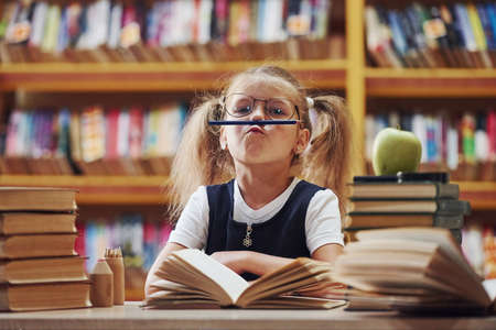 Pencil on lips. Cute little girl with pigtails is in the library. Apple on the books.の写真素材