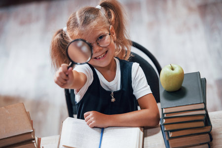 Holds magnifier. Cute little girl with pigtails is in the library. Apple on the books.