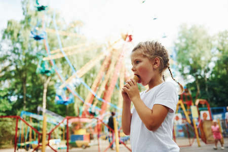 Cute little girl eats ice cream in the park at daytime near attractions.の写真素材