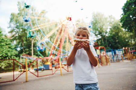 Cute little girl eats donuts in the park at daytime near attractions.の写真素材
