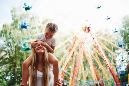 Daughter sits on the shoulders. Cheerful little girl her mother have a good time in the park together near attractions.の写真素材