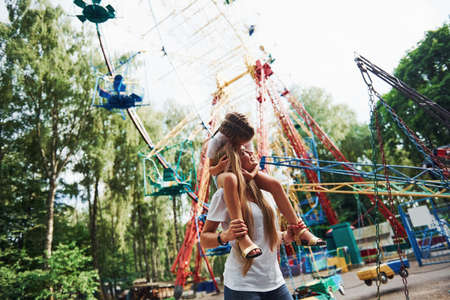 Walking with daughter on the shoulders. Cheerful little girl her mother have a good time in the park together near attractions.の写真素材