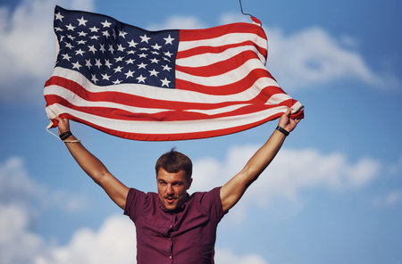 Patriotic happy man waving American Flag against cloudy blue sky.