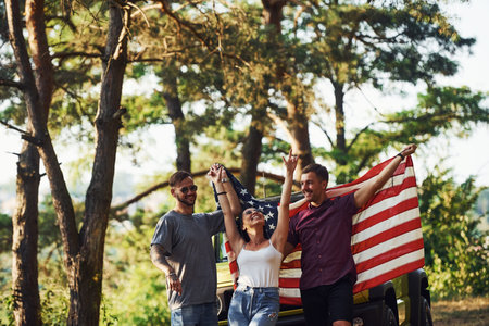 In the forest. Friends have nice weekend outdoors near theirs green car with USA flag.