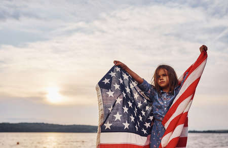 Patriotic female kid with American Flag in hands. Against cloudy sky.の写真素材