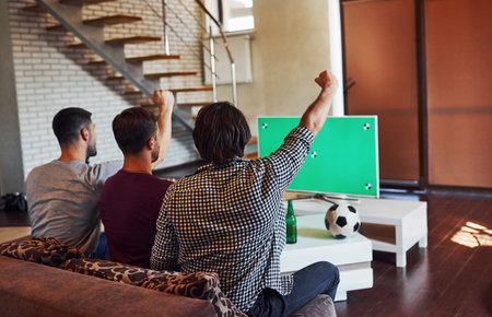 With hands up. Excited three friends watching soccer on TV at home together.