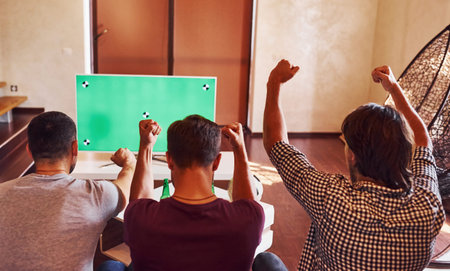 With hands up. Excited three friends watching soccer on TV at home together.