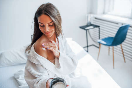 Bodycare by using cream. Portrait of beautiful brunette in white clothes that sits on the bed in room. Nice lighting.の写真素材