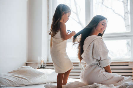 Helping to made hairstyle. Young mother with her daugher have beauty day indoors in white room.の写真素材