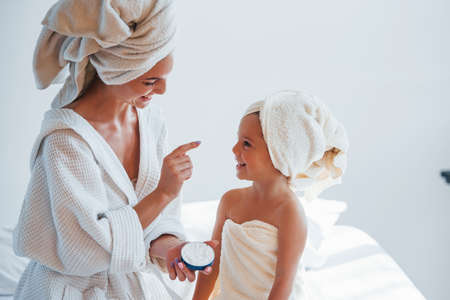 Using cream to clear skin. Young mother with her daugher have beauty day indoors in white room.の写真素材