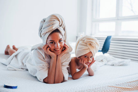 Lying down on white bed together. Young mother with her daugher have beauty day indoors in room.