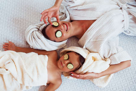 Young positive mother and daughter with towel on head lying on the bed together with cucumber.の写真素材