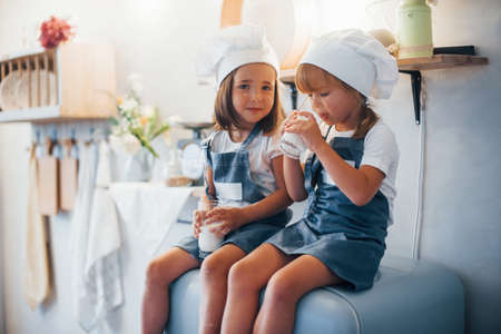 Sitting on the fridge. Family kids in white chef uniform eats food on the kitchen.の写真素材