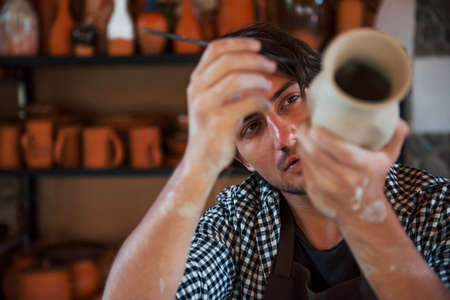 Young ceramist holds his fresh handmade pot and makes some detalizing by special tool stick.の写真素材