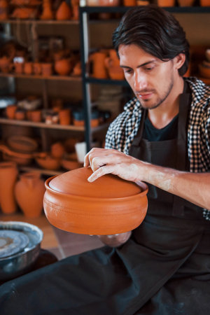 Young ceramist holds fresh handmade product made of clay in hands and looks at results of his work.