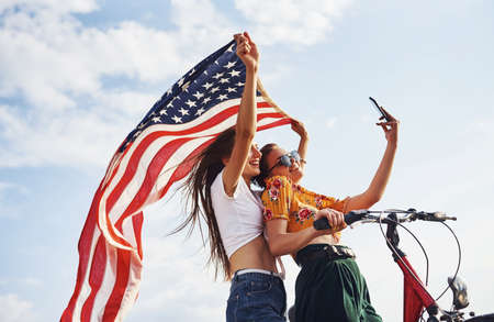 Two patriotic cheerful women with bike and USA flag in hands makes selfie.の写真素材