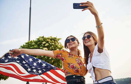 Green tree at background. Two patriotic cheerful women with USA flag in hands making selfie outdoors in park.の写真素材