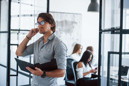 Thoughtful look. Man in glasses with notepad and pen in hands stands in the office opposite employees.の写真素材