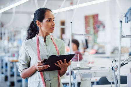 Brunette dressmaker works in the factory and holds notepad in hands.の写真素材