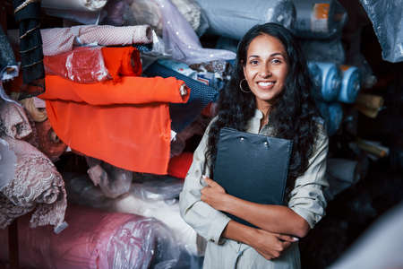 Positive smiling employee with notepad is in the storage full of cloth.の写真素材