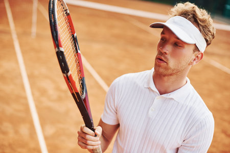 Holds racket in hands. Young tennis player in sportive clothes is on the court outdoors.