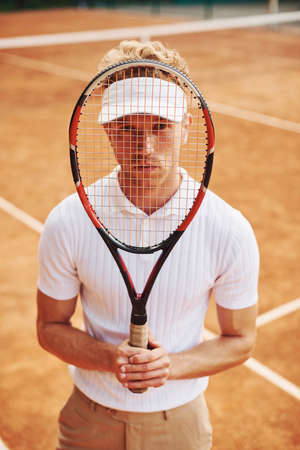 Holds racket in hands. Young tennis player in sportive clothes is on the court outdoors.の写真素材