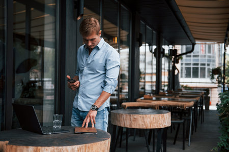 Holds phone in hand. Front view of guy in jeans that stands in the cafe at daytime.