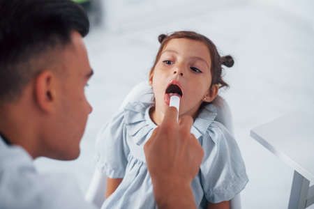 Uses tonsil to check throat. Young pediatrician works with little female visitor in the clinic.の写真素材