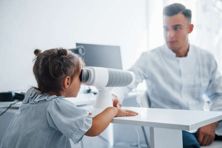 Looks into special device. Young ophthalmologist is with little female visitor in the clinic.の写真素材