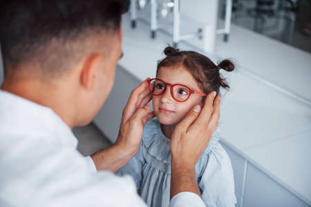 Young pediatrician in white coat helps to get new glasses for little girl.の写真素材