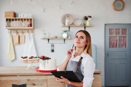 Woman with notepad in hands stands indoors at kitchen with cookies behind.の写真素材