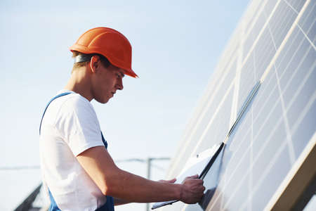 With documents. Male worker in blue uniform outdoors with solar batteries at sunny day.の写真素材