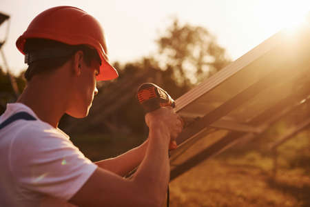 Using cordless screwdriver. Male worker in blue uniform outdoors with solar batteries at sunny day.の写真素材