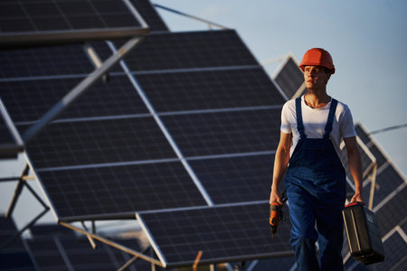 Holds case with equipment in hand. Male worker in blue uniform outdoors with solar batteries at...