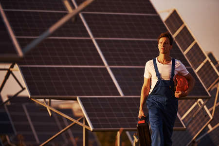 Holds case with equipment in hand. Male worker in blue uniform outdoors with solar batteries at sunny day.の写真素材