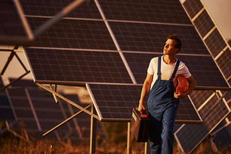 Holds case with equipment in hand. Male worker in blue uniform outdoors with solar batteries at sunny day.の写真素材